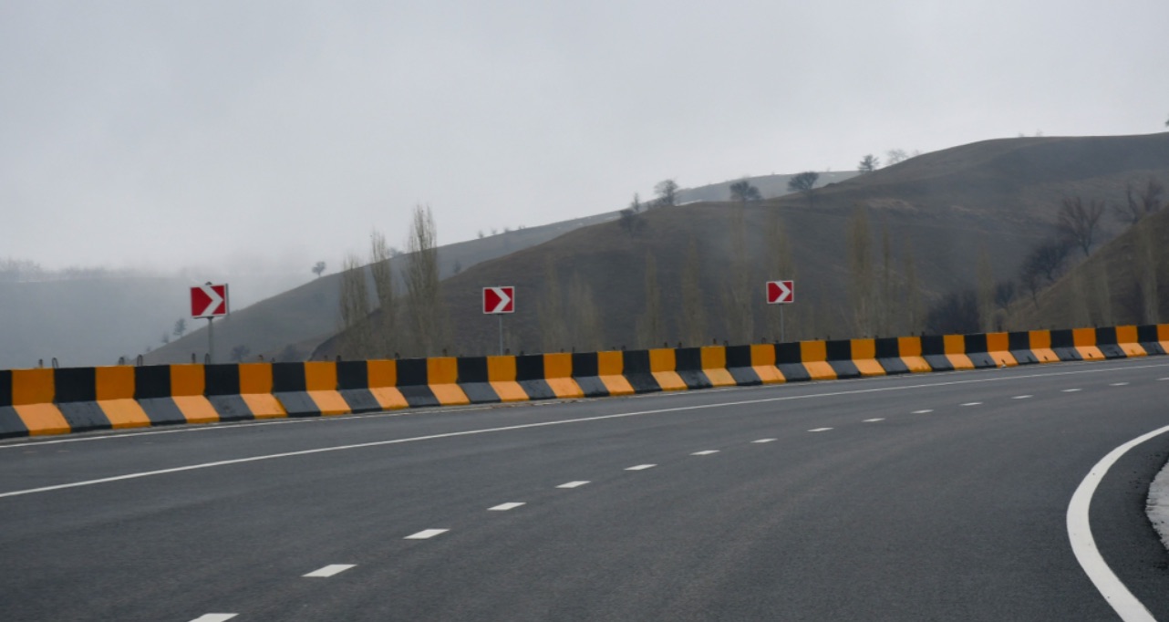 Empty road curves right past black-and-yellow barriers and chevron signs, with misty hills under a gray sky.